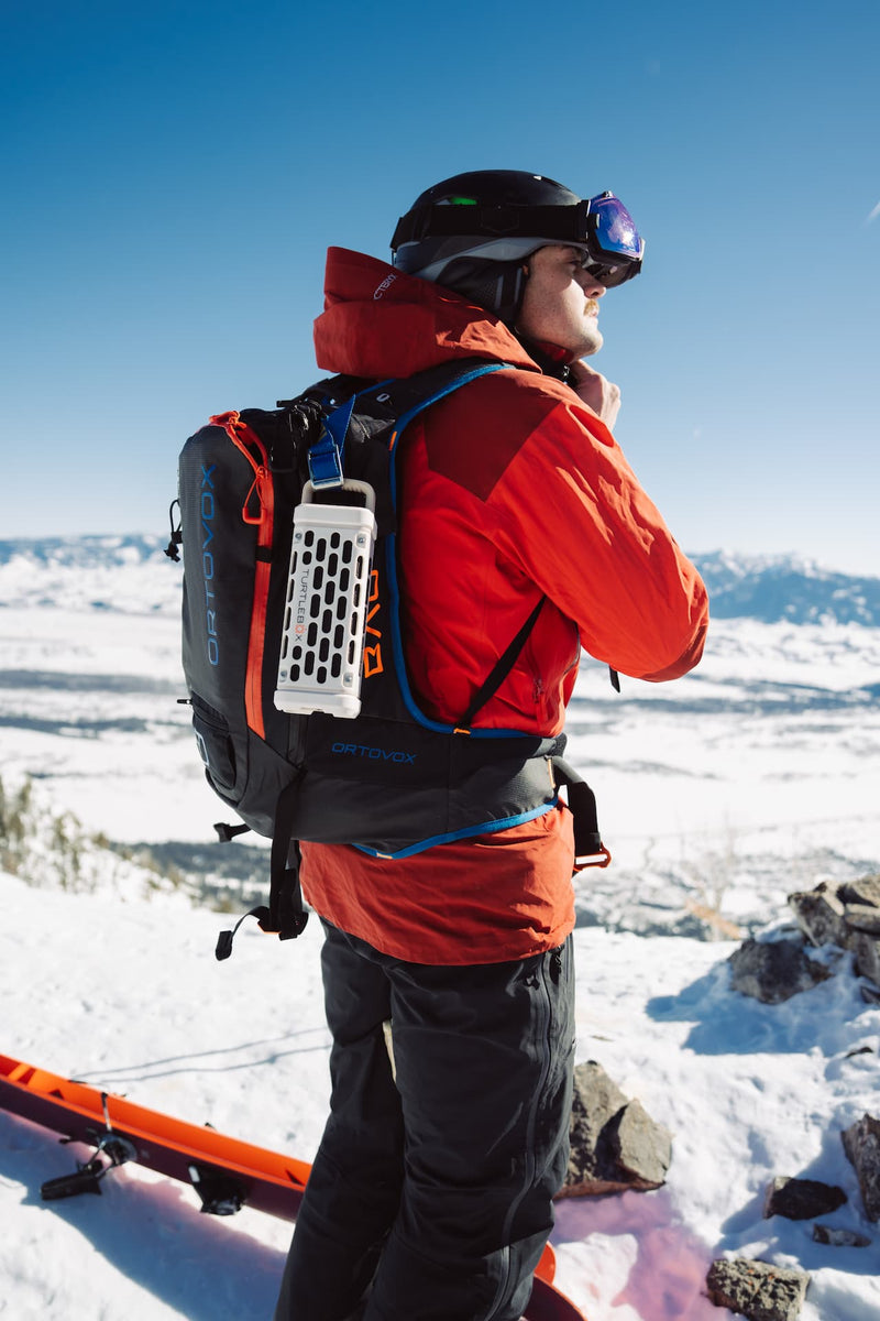 A lifestyle photo of a guy in the snow with Ranger speaker in Ivory attached to his back pack. #base-color_ivory