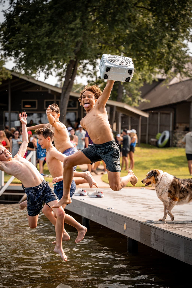 Kids jumping off a dock into a lake, one holding a white Turtlebox Original Gen 3 speaker overhead