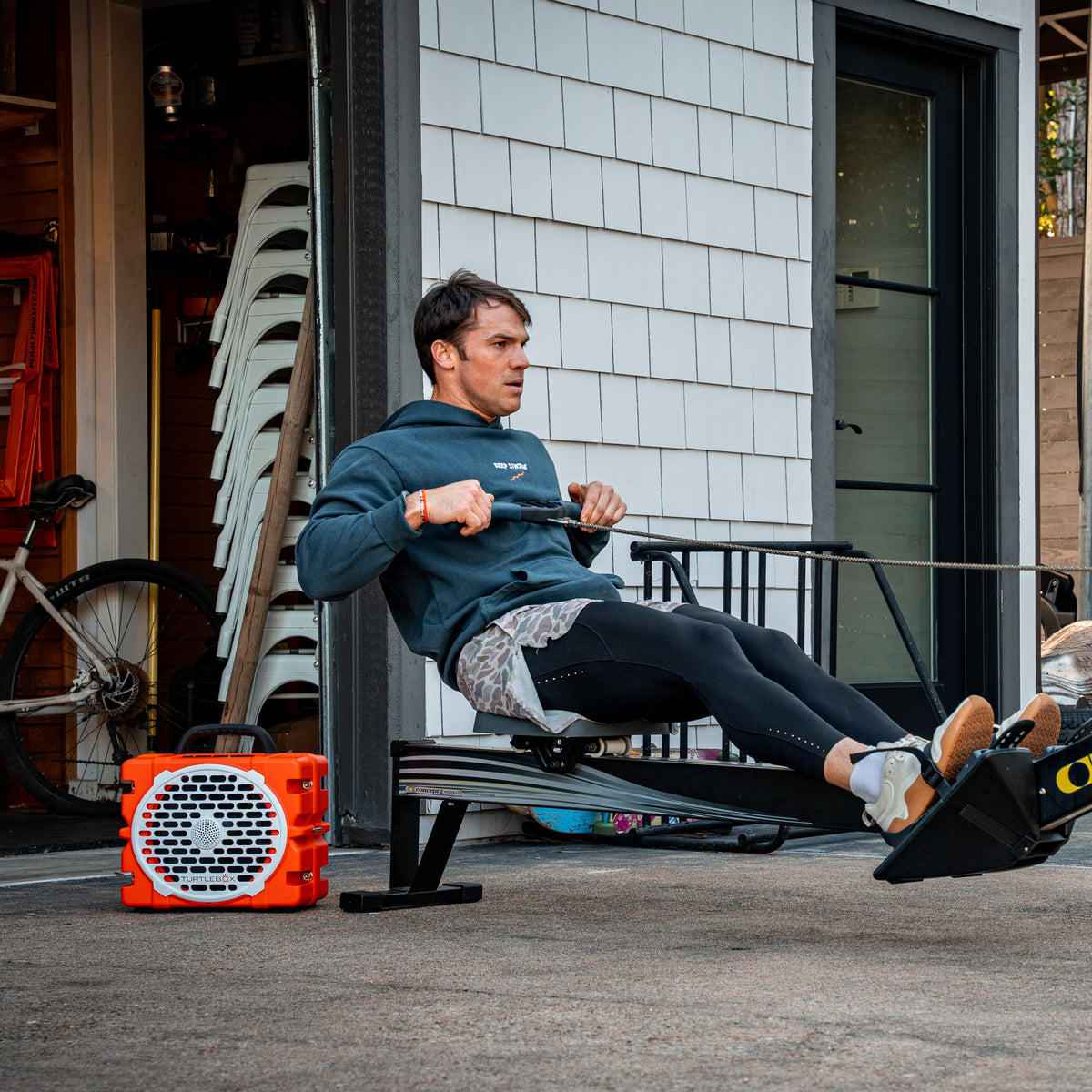 Man using a rowing machine outdoors next to an orange Grande with a building and trees in the background