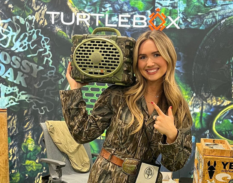 Maggie Williams, Turtlebox Ambassador ,holding a Full Foliage Speaker on her shoulder in an exhibition booth.