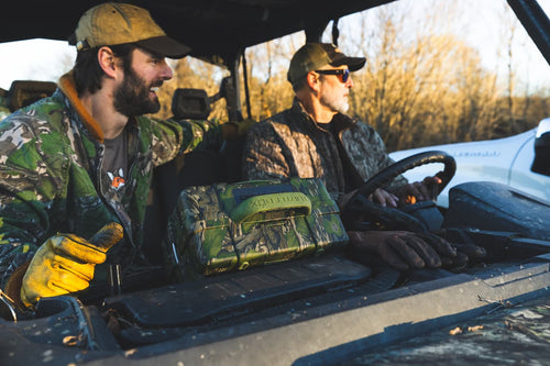 Two guys in a vehicle with Mossy Oak Full Foliage Turtlebox speaker