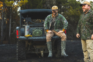 Two guys smiling on the vehicle rear listening music on their Mossy Oak Full Foliage Turtlebox speaker