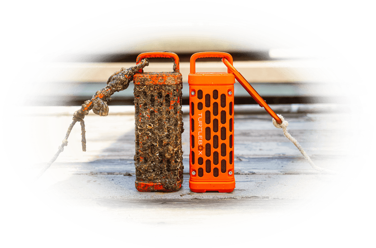 Two orange waterproof speaker-style devices with carrying handles and cords, shown side by side on a white surface with a blurred outdoor background visible through an oval reflection effect.