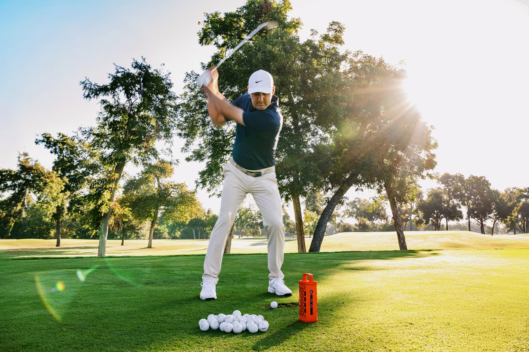 A lifestyle photo from gplfer Scottie Sheffler taking a swing on a golf course with trees in the background #base-color_orange