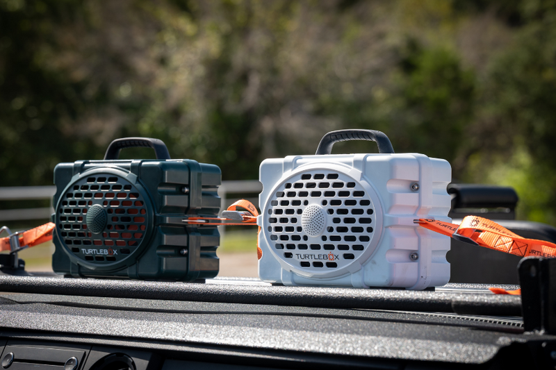 lifestyle photo of a green turtlebox and a white turtlebox strapped to a vehicle with orange Turtlebox straps